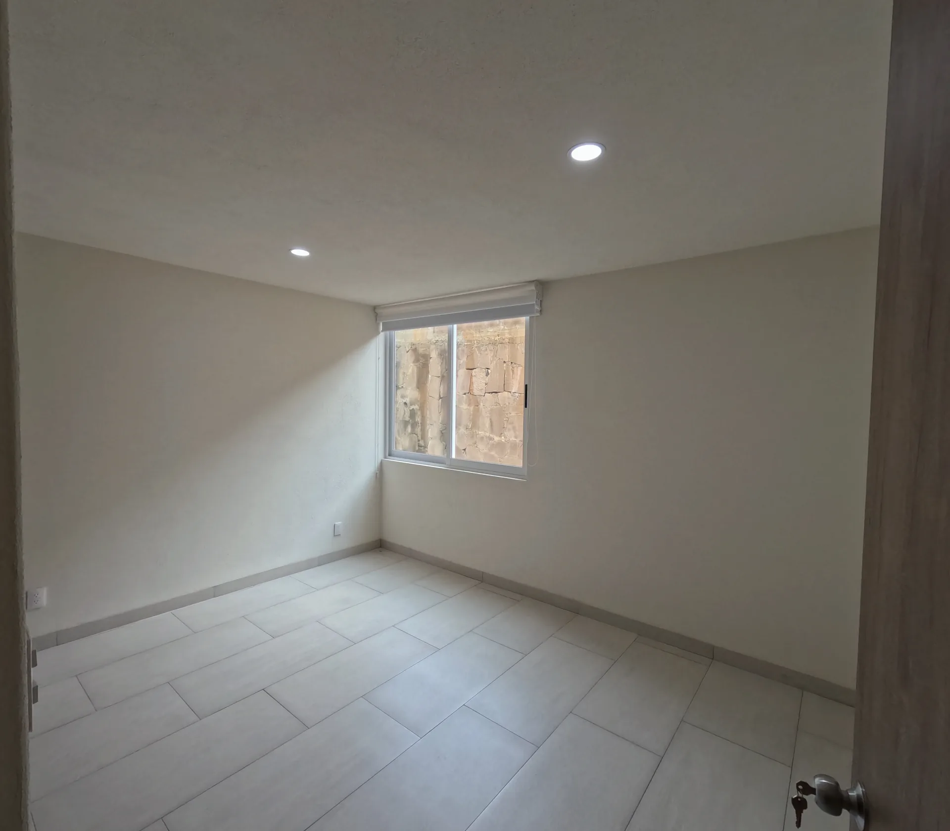 Recamara con ventana de aluminio, piso de loseta y vista hacia pared de piedra del terreno colindante.
