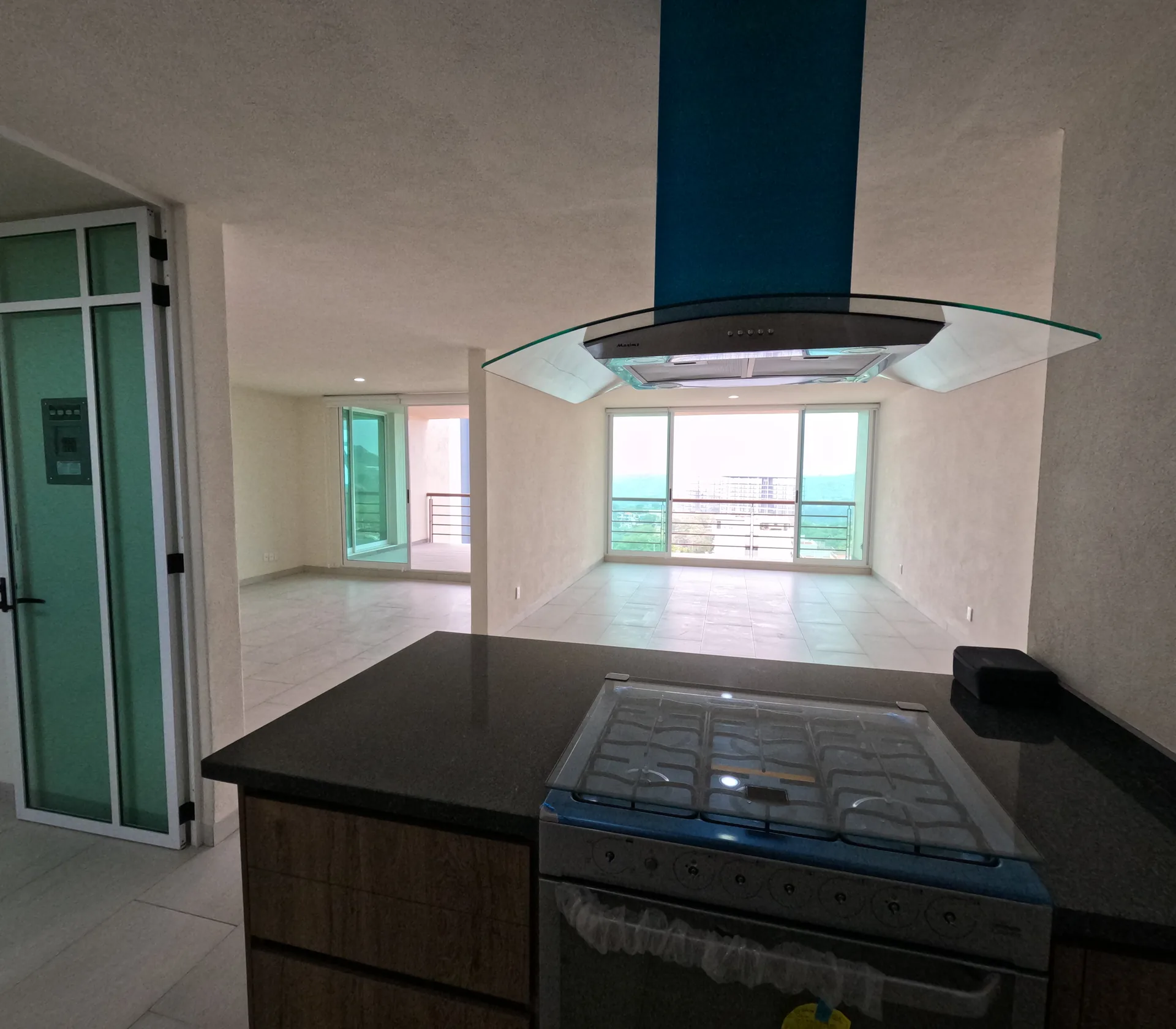 Cocina de departamento tipo con isla central, estufa de gas y campana extractora; al fondo se ve la sala con balcon.