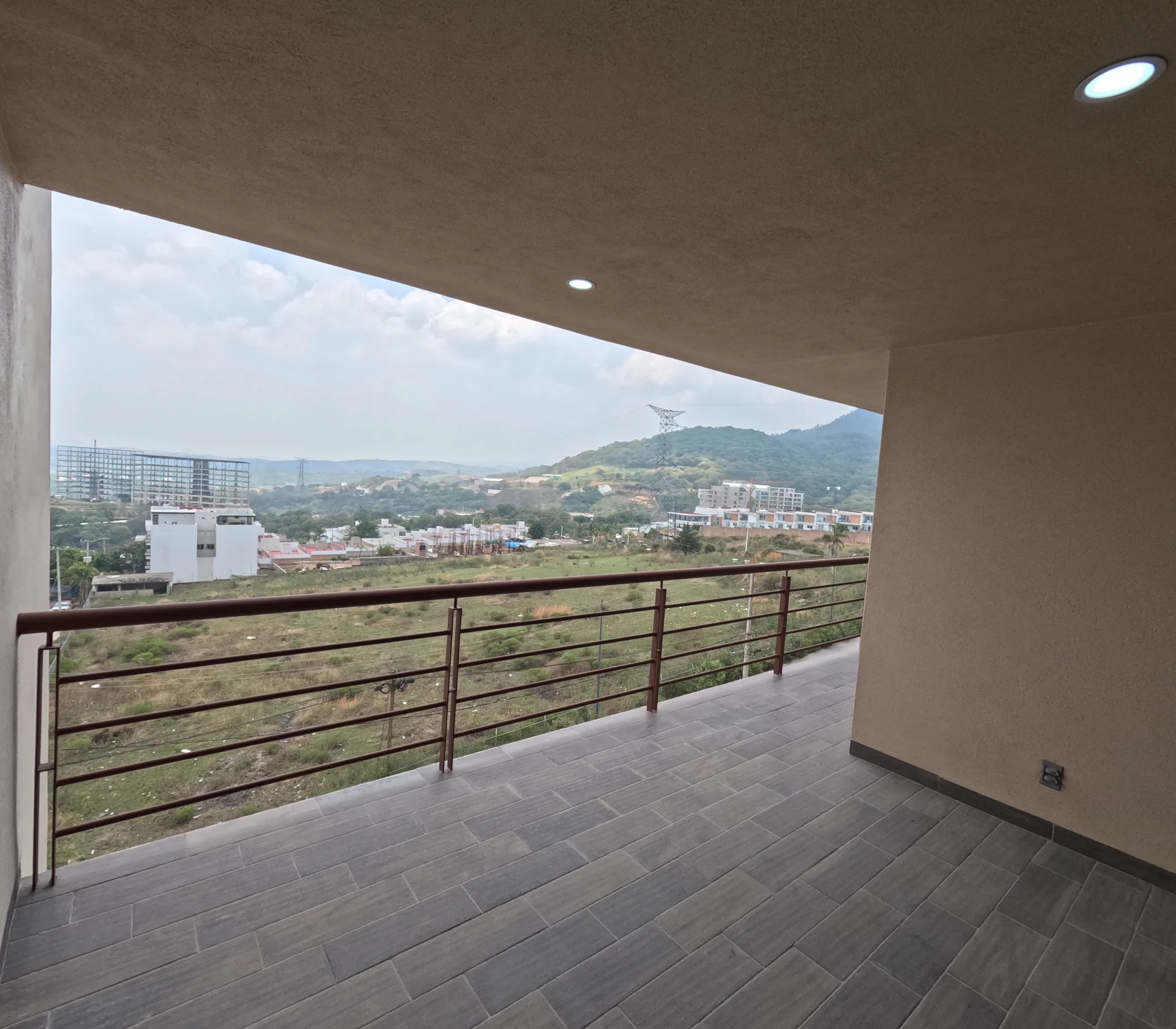 Balcon-terraza con piso gris y barandal metalico rojo, vista abierta al entorno urbano y montana de fondo.